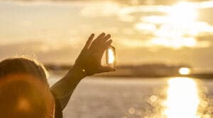 A hand holds a crystal up to the sunrise over a calm sea, capturing the light and creating a serene, reflective atmosphere.