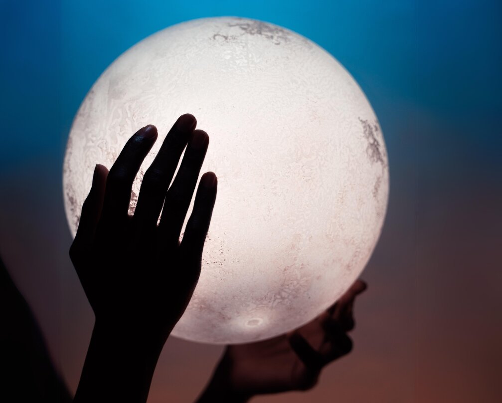 A hand silhouetted against a glowing white orb resembling the moon, set against a blue and orange gradient background.