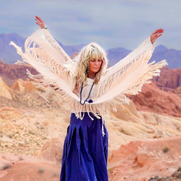 Woman raising arms in a desert landscape, wearing a fringed shawl and blue skirt, evoking a sense of freedom and tranquility.