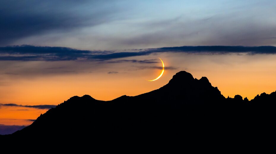 Crescent moon setting behind dark mountain silhouette at sunset, with a vibrant sky transitioning from orange to blue.