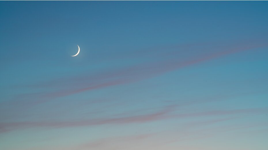 Crescent moon in a clear blue sky at dusk, with soft pink clouds, symbolizing tranquility and peace.