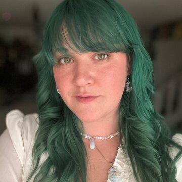 Portrait of a woman with green hair, wearing a white top and jewelry, smiling softly against a neutral background.