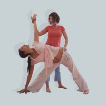Two women practicing yoga together; one demonstrating a pose while the other offers support, emphasizing connection and guidance.